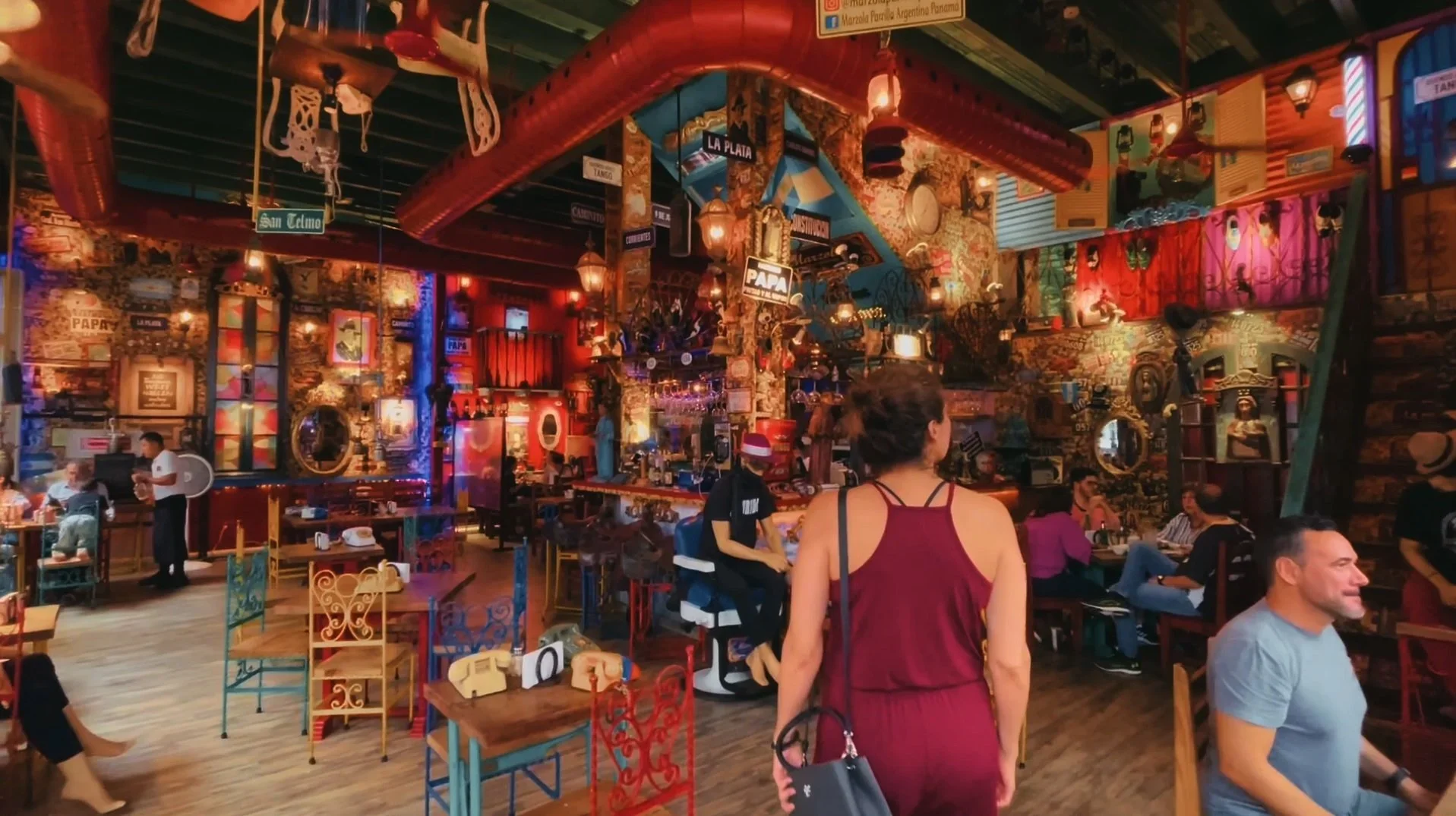 Interior of a lively restaurant decorated with colorful, eclectic artwork and ornaments, featuring patrons seated at tables and a woman with a ponytail in a red dress walking through.