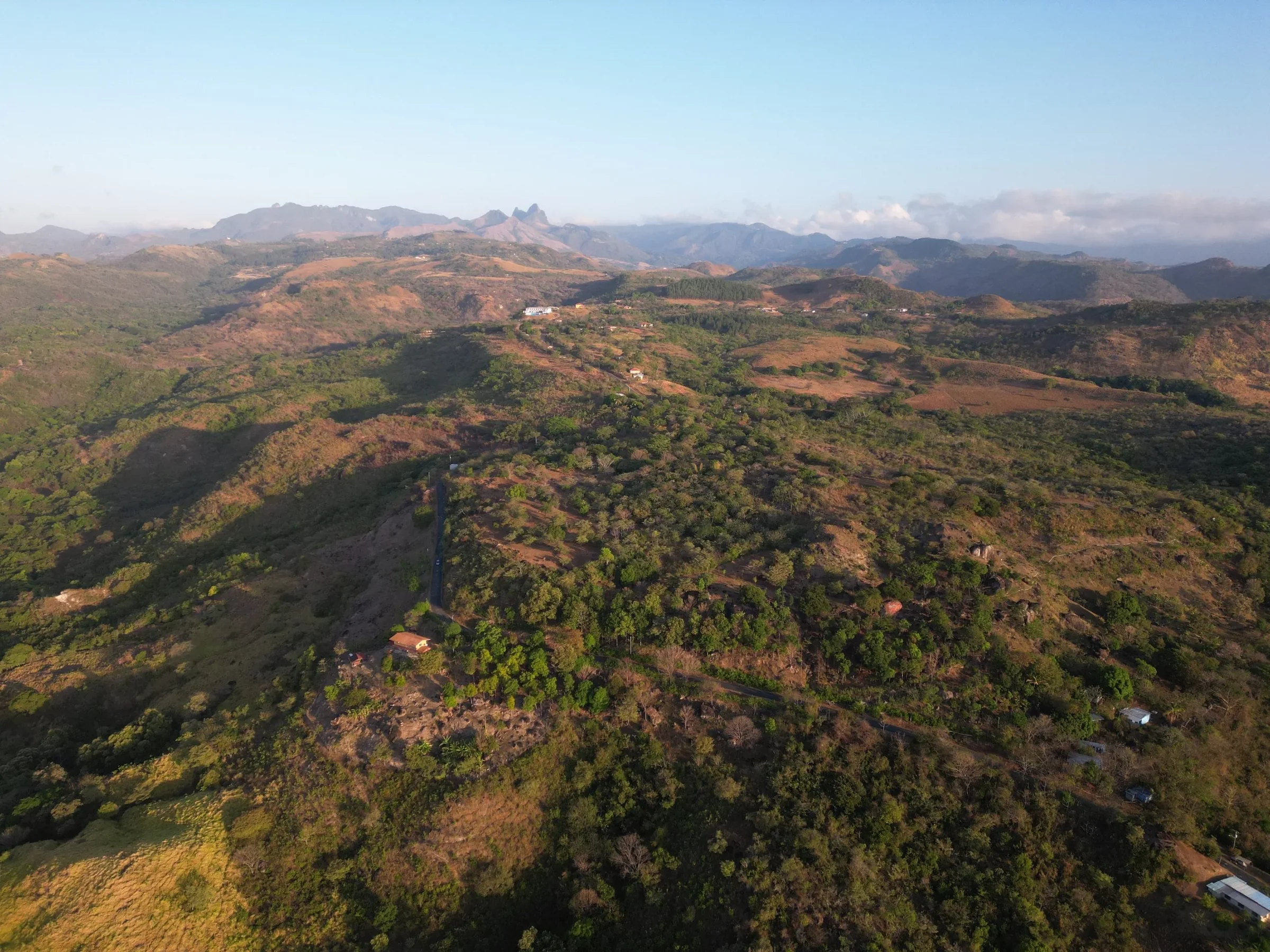 Aerial view of rolling green and brown hills with scattered trees, small houses, and distant mountains under a clear blue sky.