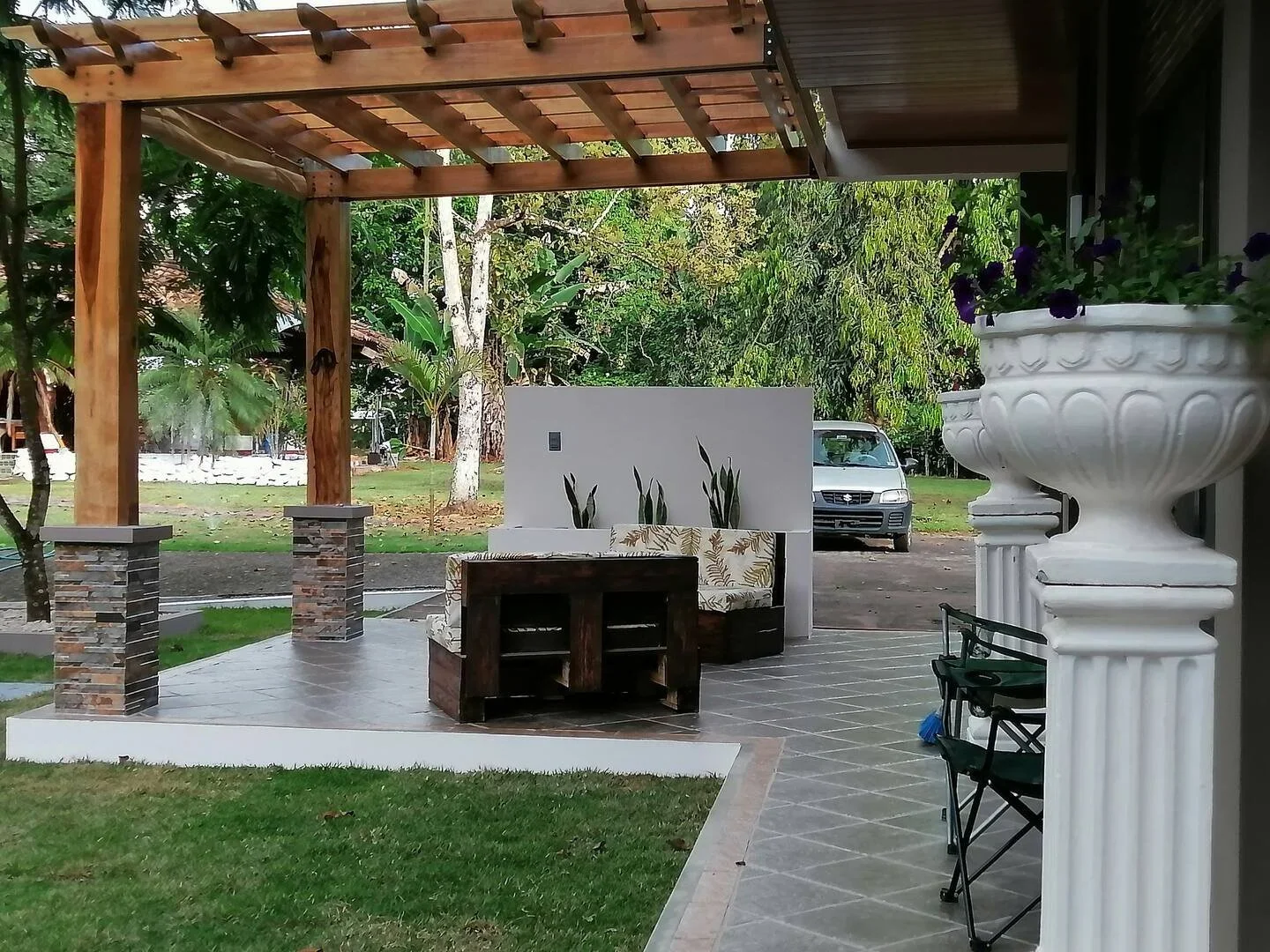 A patio area with a wooden pergola, brick pillars, and outdoor seating including a bench with floral cushions. There are large potted plants and trees in the background, with a car parked nearby.