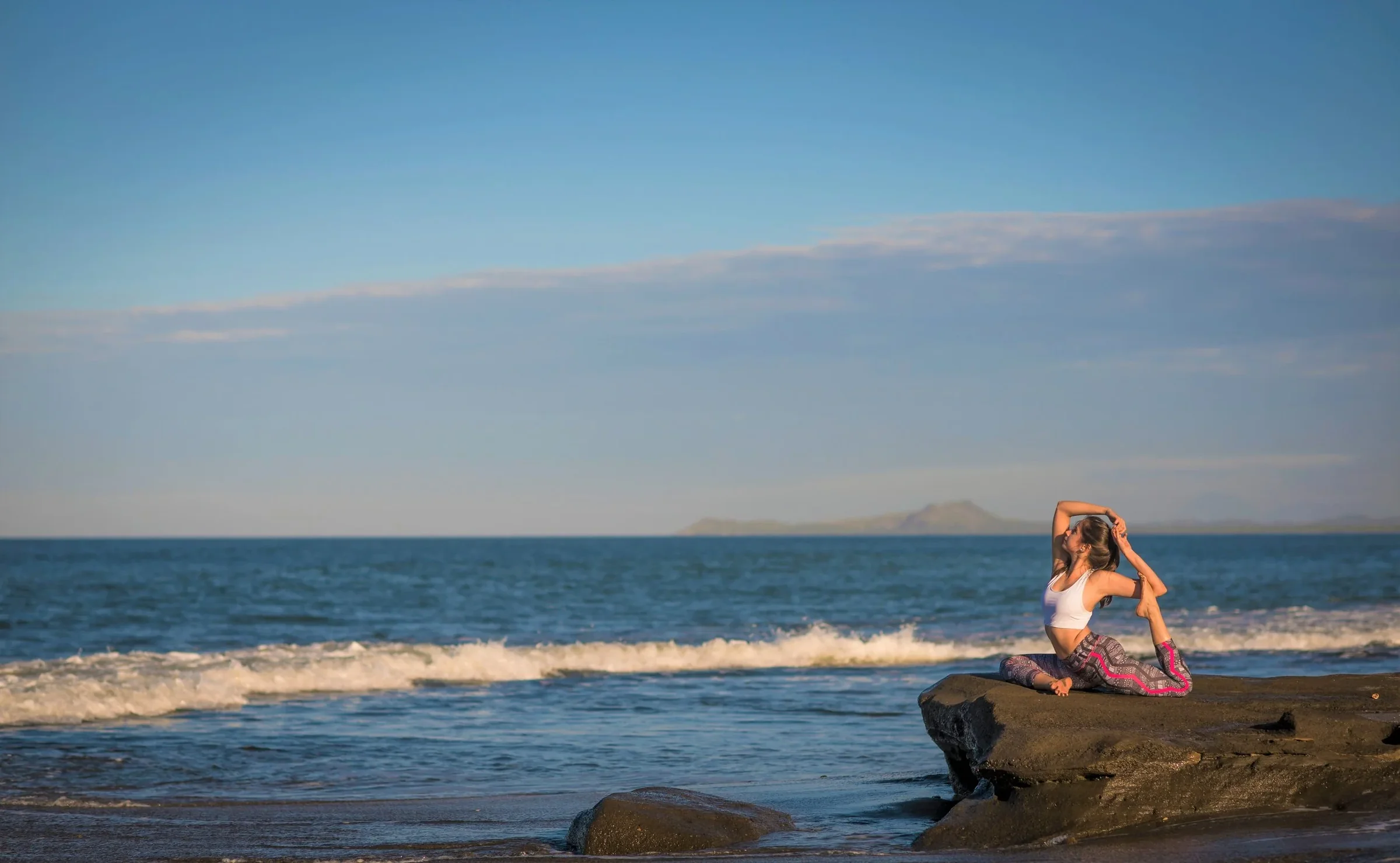A woman practicing yoga on a rocky beach at sunset, with the ocean and distant islands in the background.