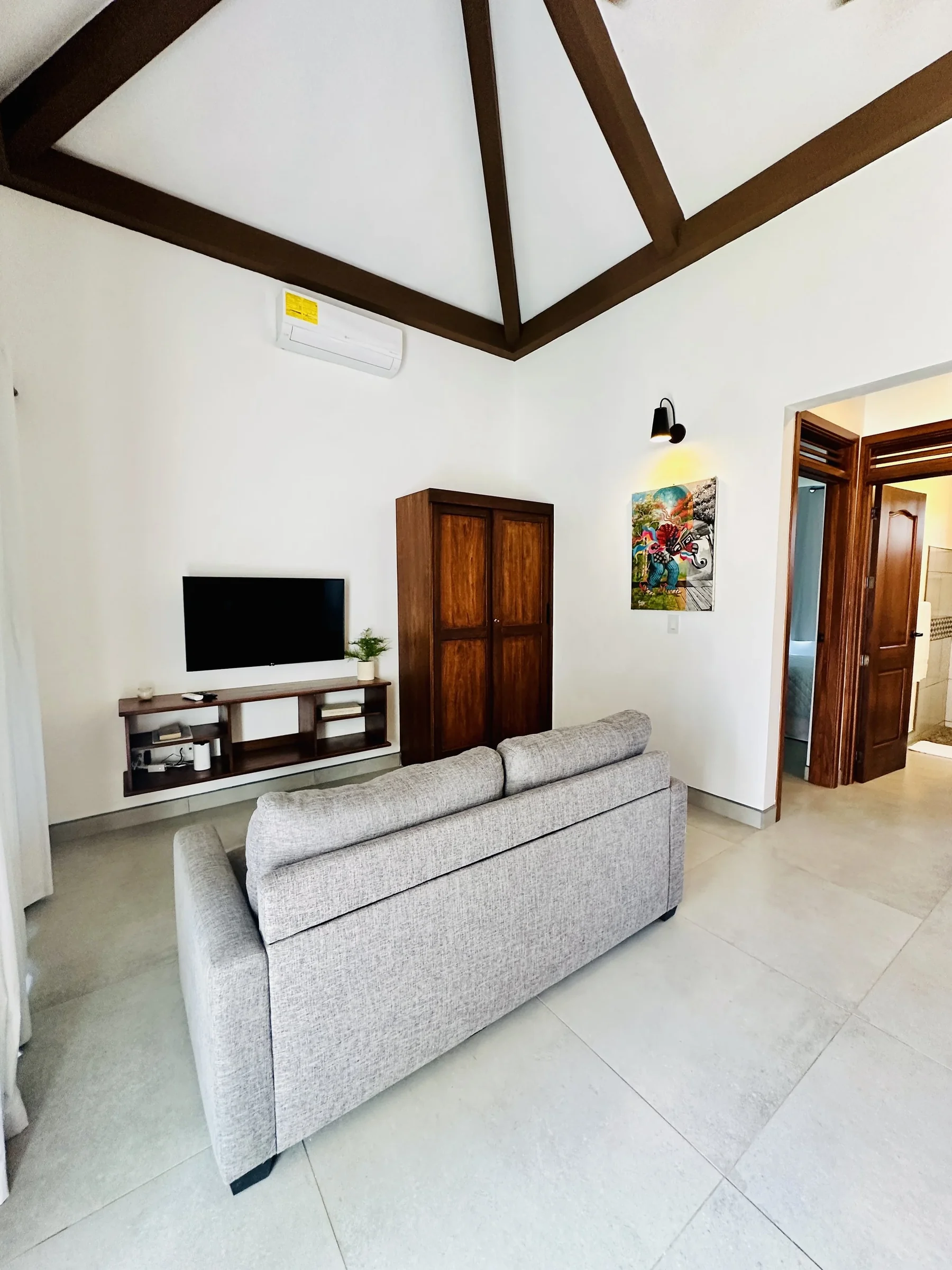 Living room with a beige sofa, wall-mounted TV, wooden cabinet, colorful painting, and doorway leading to bathroom.