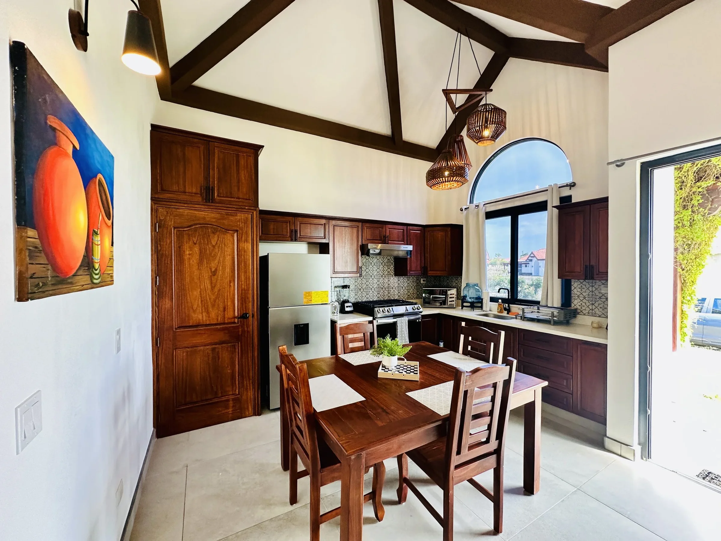 A bright kitchen with wooden cabinets, a dining table with four chairs, a stainless steel refrigerator, a stove, and a window with a view outside. Woven hanging pendant lights and artwork on the wall add decor.