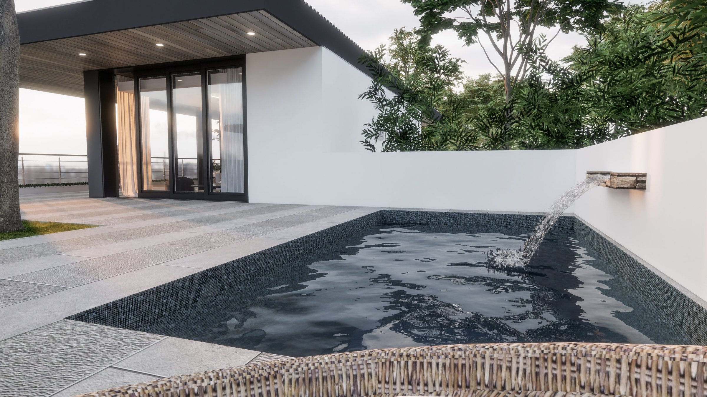Modern backyard with a small rectangular water fountain with a slow waterfall feature, tiled black lining, and surrounded by a paved patio, a white wall with greenery behind, and a house with large glass doors and wooden ceiling.
